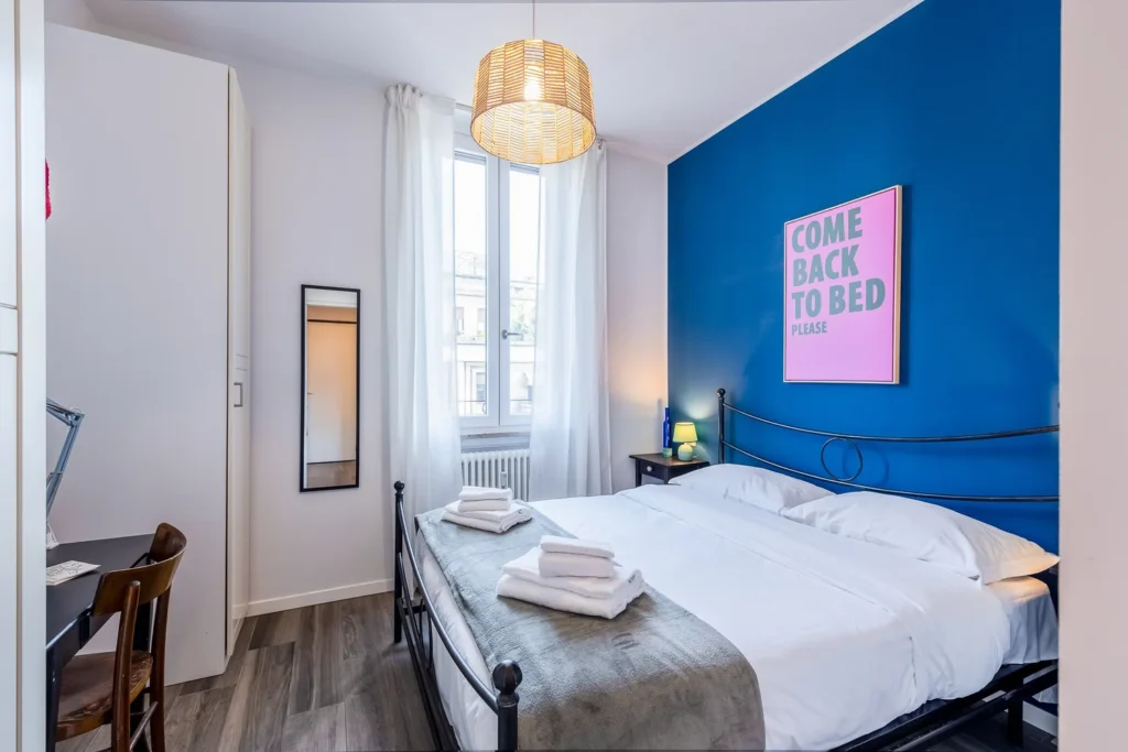 Bright bedroom with blue accent wall, double bed, and tall window in the Colorful Eclectic apartment by Townhomes Milano.