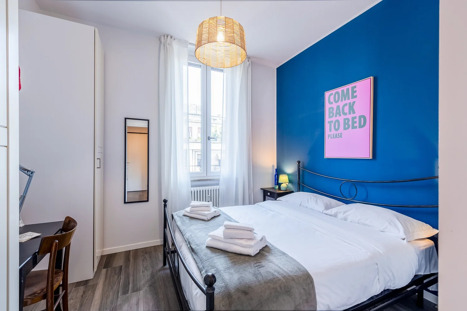 Bright bedroom with blue accent wall, double bed, and tall window in the Colorful Eclectic apartment by Townhomes Milano.