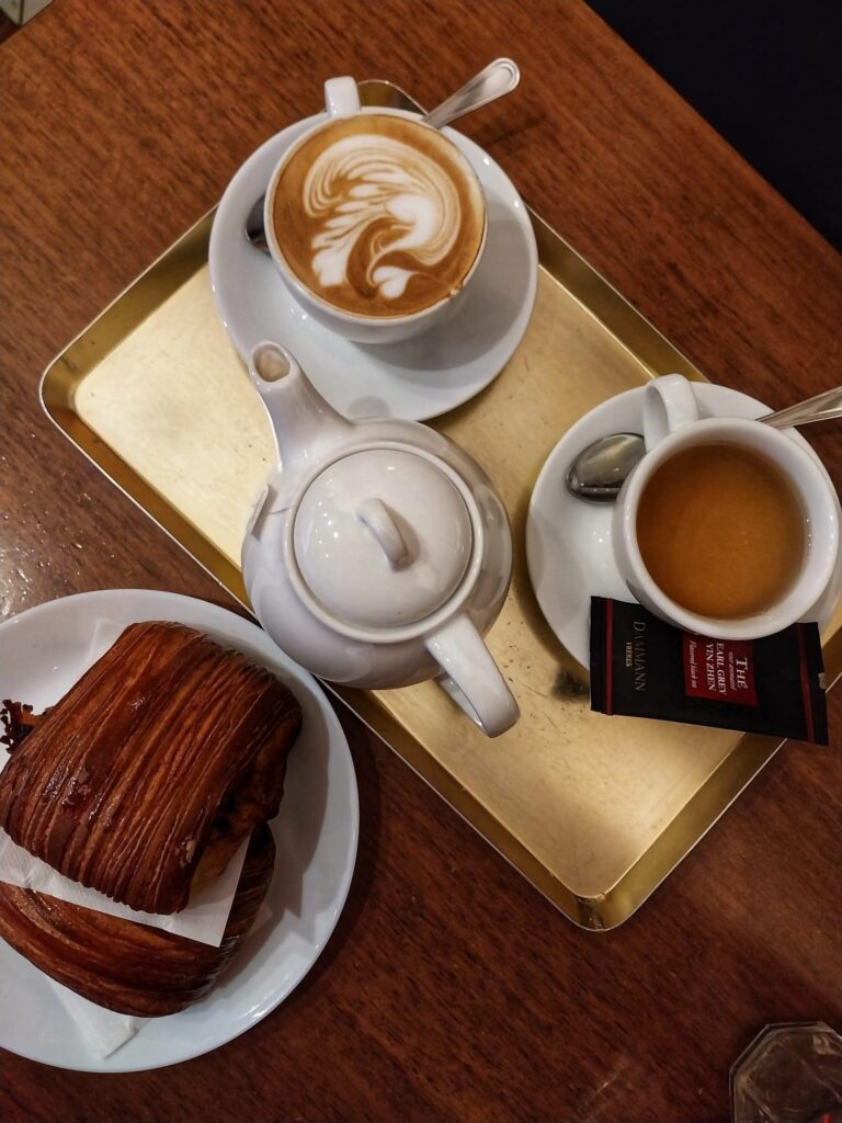 An overhead view of a latte with heart art, a black coffee, and a chocolate croissant on a gold tray over a dark wood table.