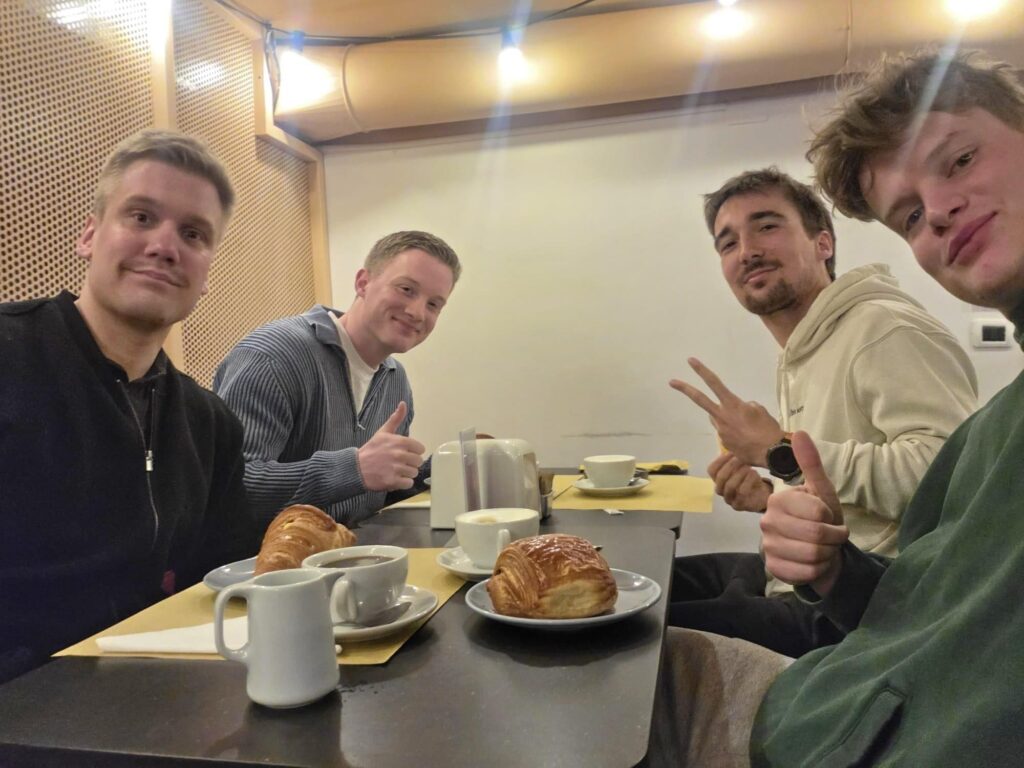 Four men smiling at a cafe table for breakfast at Pave, Milan. The group is enjoying Italian coffee and artisanal pastries in a warm, informal indoor setting on Via Felice Casati.