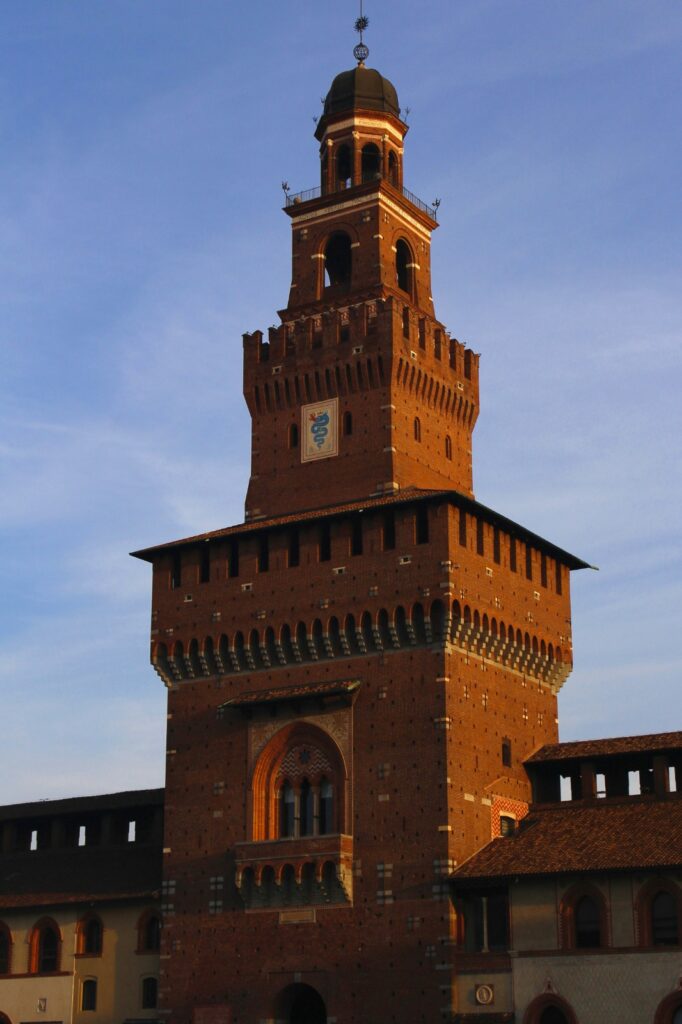The historic Filarete Tower of Castello Sforzesco in Milan against a blue sky, representing the 14th-century red-brick fortress and its surrounding cultural parkland.