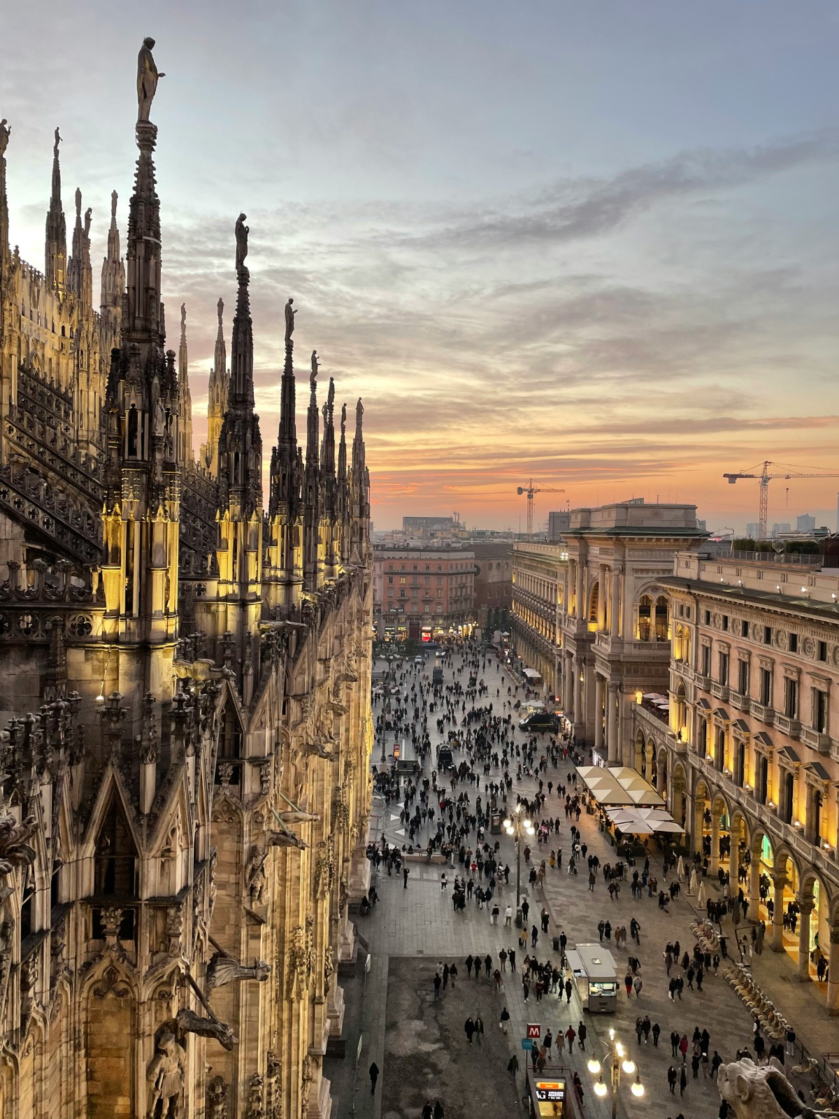 Panoramic city view from the Duomo di Milano terraces at sunset, highlighting the intricate Gothic marble spires and urban skyline.