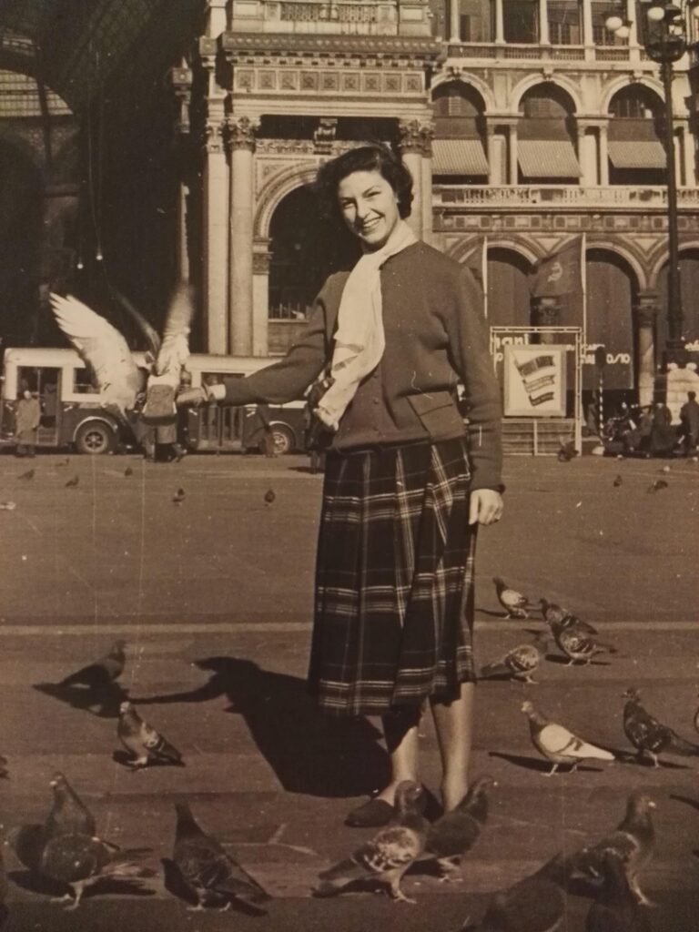 Vintage black-and-white photo of a woman with pigeons in front of Galleria Vittorio Emanuele II, capturing the timeless local atmosphere of Milan's living room.