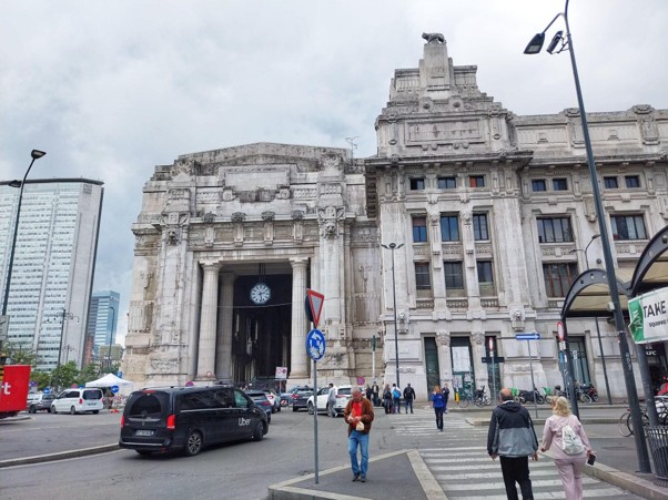 Historic Milan landmark with ornate stone architecture, clock tower, and busy street crossing